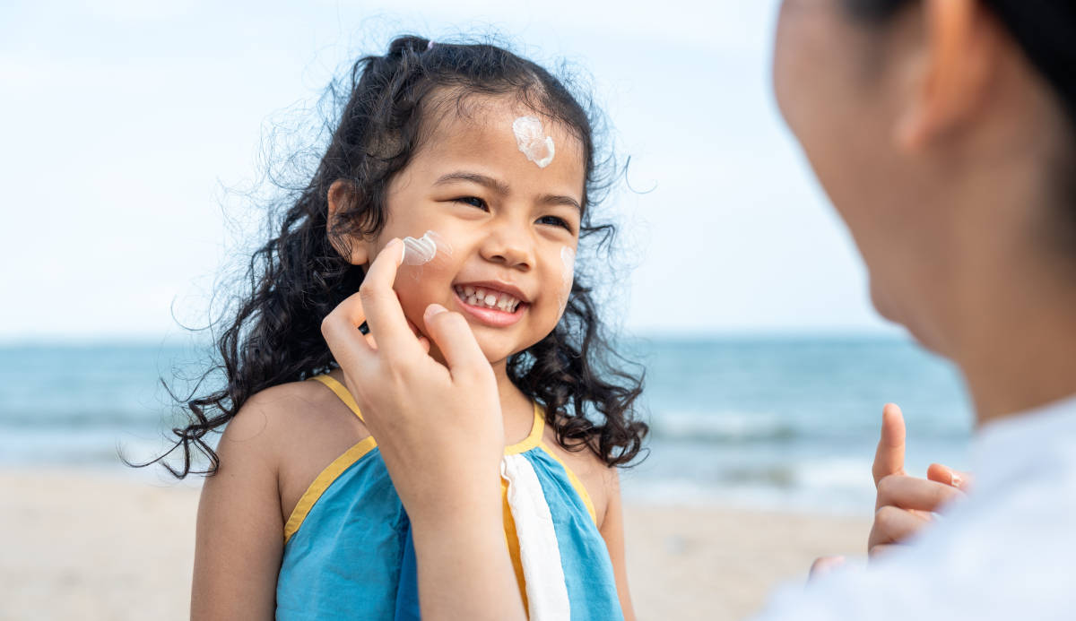 Seorang ibu sedang mengoleskan sunscreen saat anak bermain di pantai